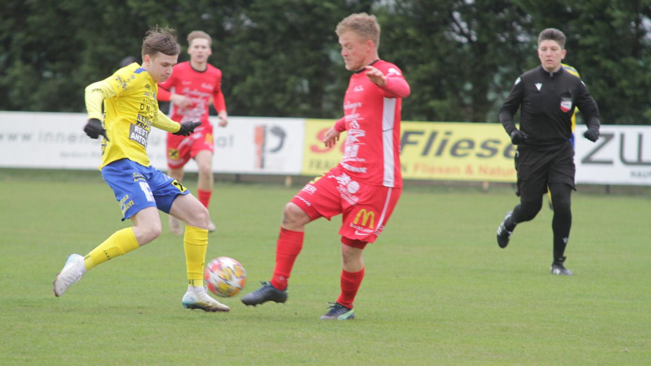Three soccer players are on a field, two in red uniforms, one in yellow. The player in yellow is kicking the ball. Behind them is a banner with the word Flies.