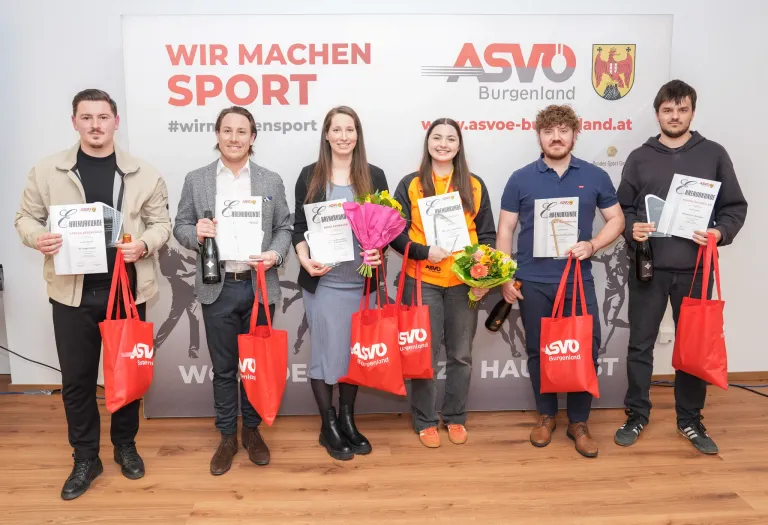 A group of people standing in front of a banner that says 'Wir machen sport' and holding certificates. They are smiling and holding red bags and bottles. They are wearing different colored outfits and shoes.