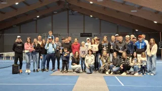Eine Gruppe von Menschen posiert für ein Foto auf einem Tennisplatz. Einige von ihnen halten Trophäen. Hinter ihnen hängen Banner an der Wand.