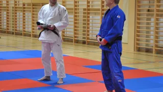 Zwei Männer in Kampfsportuniformen stehen auf einem blau-roten Mattenboden in einer Sporthalle, möglicherweise bereit für einen Wettkampf. Einer hält eine Mappe, und der andere schaut zu.