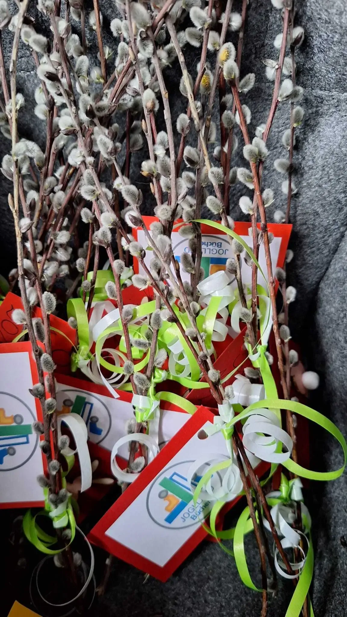 Ein Strauß Weidenzweige mit weißen Blüten, die mit grünen und weißen Bändern gebunden sind. Die Zweige sind in einer roten und weißen Karte mit einem Logo.