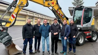 Fünf Männer posieren vor einem großen gelben Baufahrzeug, möglicherweise einem Bulldozer, auf einer Betonoberfläche. Sie tragen Winterkleidung.