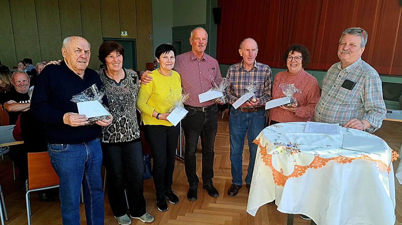Sechs Menschen stehen in einem Raum, halten Papiere und Blumen in Plastik. Zwei Männer und vier Frauen, lächelnd, einer der Männer hält eine Frau an der Taille. Dahinter steht ein Tisch mit einer Tischdecke und einem roten Vorhang.