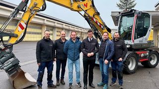 Fünf Männer stehen vor einem gelben Bagger auf einem Parkplatz. Sie alle lächeln und scheinen für ein Foto zu posieren.