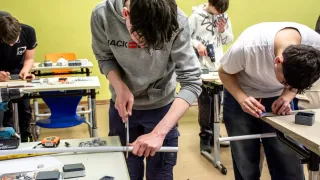 Ein Schüler in einer grauen Kapuzenjacke arbeitet mit einem Metallstab und Werkzeugen auf einem Tisch. Hinter ihm benutzt ein anderer Schüler eine Bohrmaschine. Der Klassenraum ist gelb gestrichen mit verschiedenen Werkzeugen und Tischen.