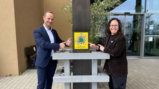 Zwei Personen, ein Mann und eine Frau, stehen neben einem Pfosten und weisen auf ein gelbes Schild mit einem grünen Kreis. Sie lächeln beide und scheinen sich in einer professionellen Umgebung zu befinden.