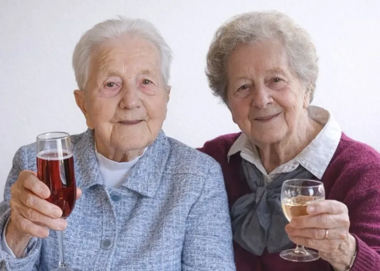 Two elderly women sitting next to each other, one holding a glass of red wine and the other a glass of white wine, smiling for the camera.