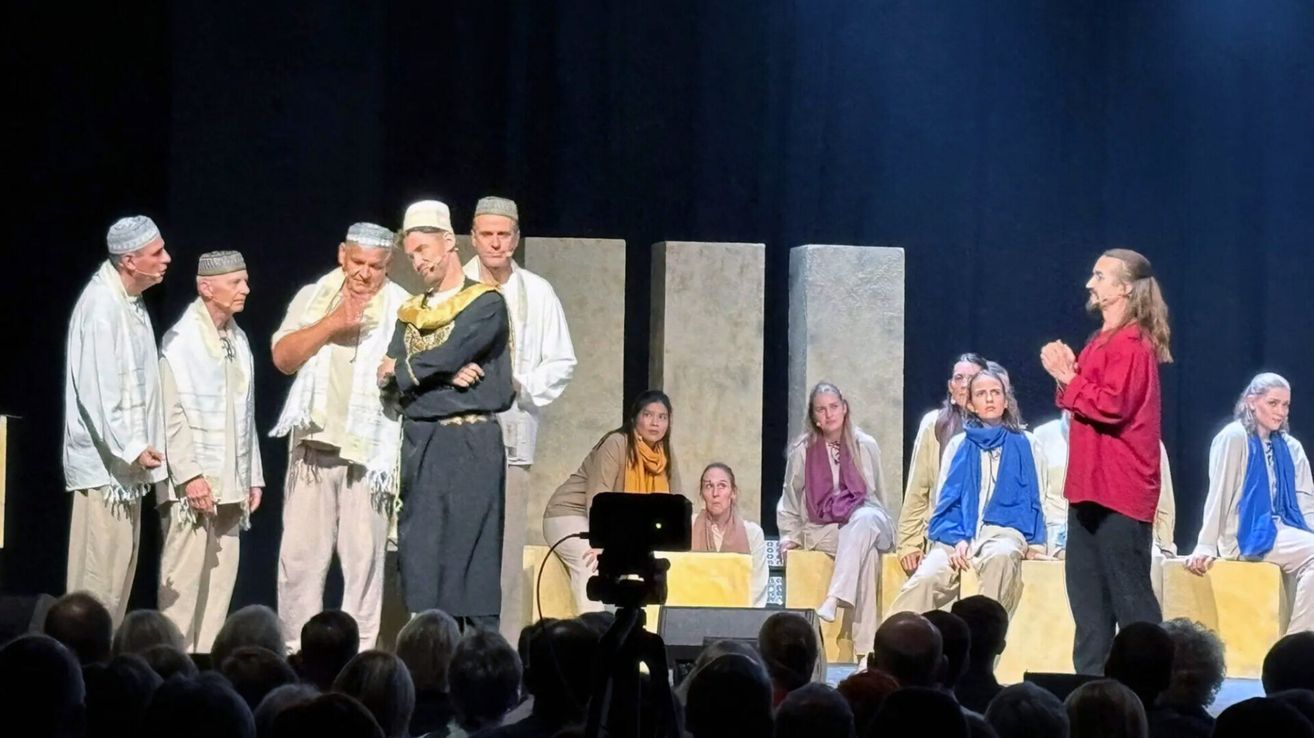 Eine Gruppe von Darstellern auf der Bühne, einige in traditioneller Kleidung, andere mit Schals, einem Hintergrund aus großen Steinsäulen, einer Kamera und einem Publikum, das zusieht.