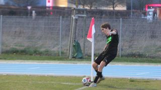 Ein Fußballspieler tritt einen Ball auf einem Feld mit einer rot-weißen Fahne neben einer Laufbahn.
