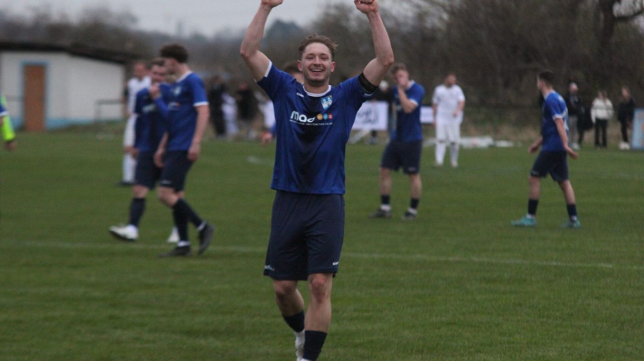 Ein Fußballspieler in einem blauen Trikot feiert einen Sieg auf einem grünen Feld mit Teamkollegen und unscharfen Zuschauern im Hintergrund.