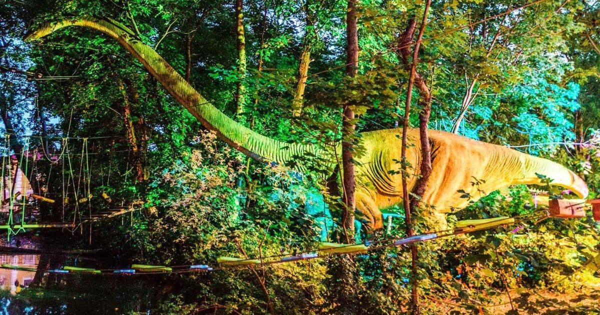 Ein Dinosaurier in einem Wald, beleuchtet mit grünen und gelben Lichtern. Bäume umgeben den Dinosaurier und werfen Schatten.