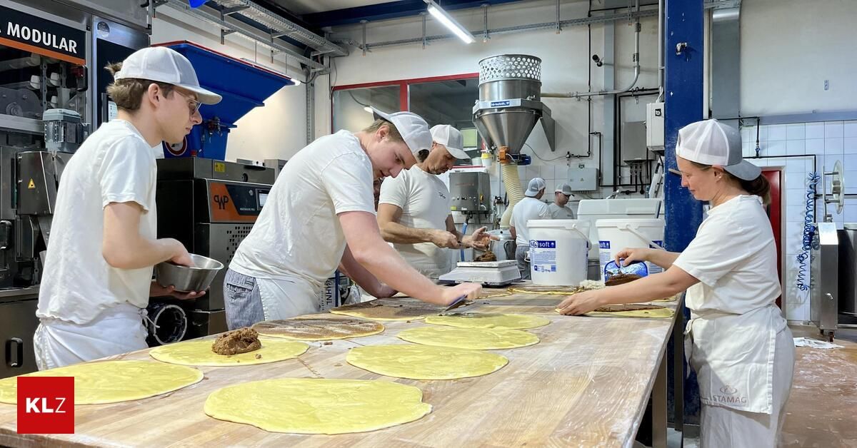 Eine Gruppe von Arbeitern in weißen Schürzen und Mützen arbeitet an einem großen Tisch voller Teig. Sie befinden sich in einer gewerblichen Küche mit industriellen Geräten im Hintergrund.