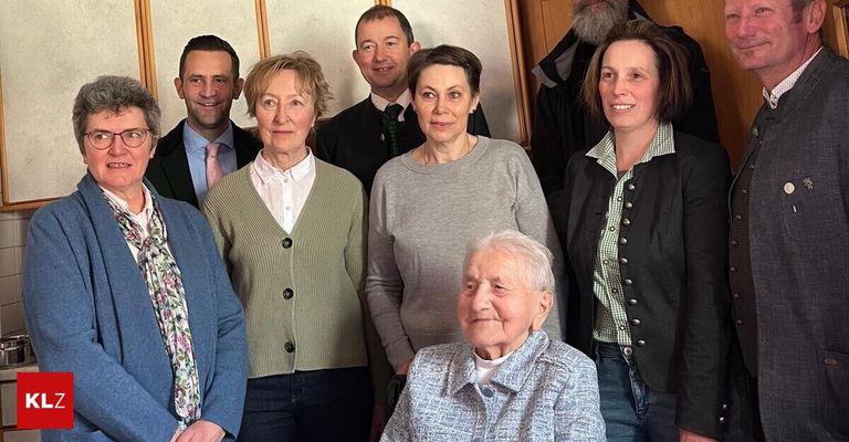A group of people are posing for a photo in a room. An elderly woman in the middle is sitting. Behind her, three women and two men are standing. They are all smiling.
