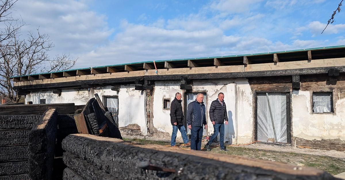 Drei Männer in Jacken inspizieren ein beschädigtes Gebäude mit beschädigtem Dach und verkohlten Trümmern. Der Himmel ist blau mit Wolken.