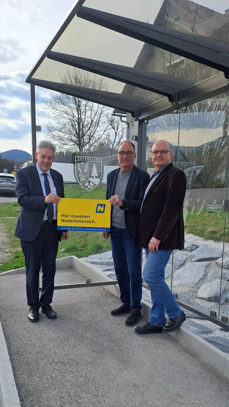 Three men stand in front of a bus shelter, holding a yellow sign with the words 'Here invests Niederosterreich'. They seem to be celebrating. Behind them is a fence with a logo and some trees.