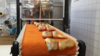 Ein Förderband transportiert mehrere golden verpackte Gegenstände durch eine Fabrik. Die Gegenstände sind orangefarben.