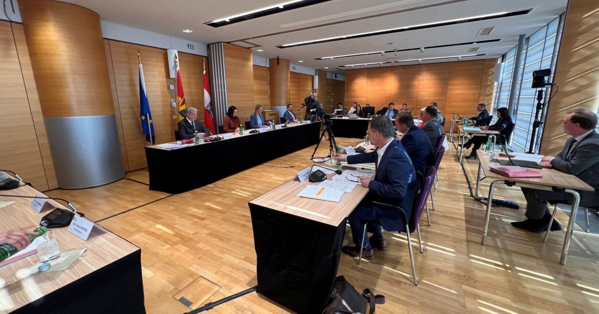 Eine Sitzung findet in einem Konferenzraum mit Holzboden statt. Die Menschen sitzen an langen Tischen mit Mikrofonen, Papieren und Flaschen. Fahnen schmücken die Wände.