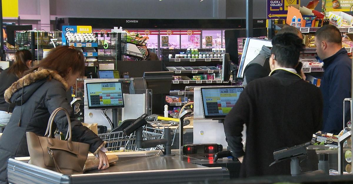 In einem Supermarkt stehen eine Frau und ein Mann an einer Kasse mit Monitoren und einem Einkaufswagen. Hinter ihnen sind Regale mit verschiedenen Produkten zu sehen.