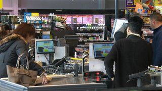 In einem Supermarkt stehen eine Frau und ein Mann an einer Kasse mit Monitoren und einem Einkaufswagen. Hinter ihnen sind Regale mit verschiedenen Produkten zu sehen.