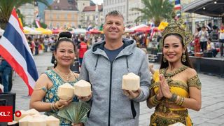 Ein Mann in einer grauen Kapuzenjacke hält Kokosnüsse mit zwei Frauen in traditioneller thailändischer Kleidung auf einem Markt.