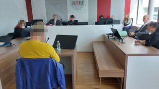 Ein Gerichtssaal mit einem Richter und einer Person am Schreibtisch. Eine Person in einem gelben Shirt sitzt auf einem Stuhl. Die Wand hat ein Logo mit dem Text 'JUSTIZ'.