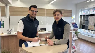 Zwei Männer in Westen stehen an einem Tisch und unterzeichnen Papiere. Einer trägt eine Brille, der andere eine Uhr. Im Hintergrund sind ein Schreibtisch und ein Heizkörper zu sehen.