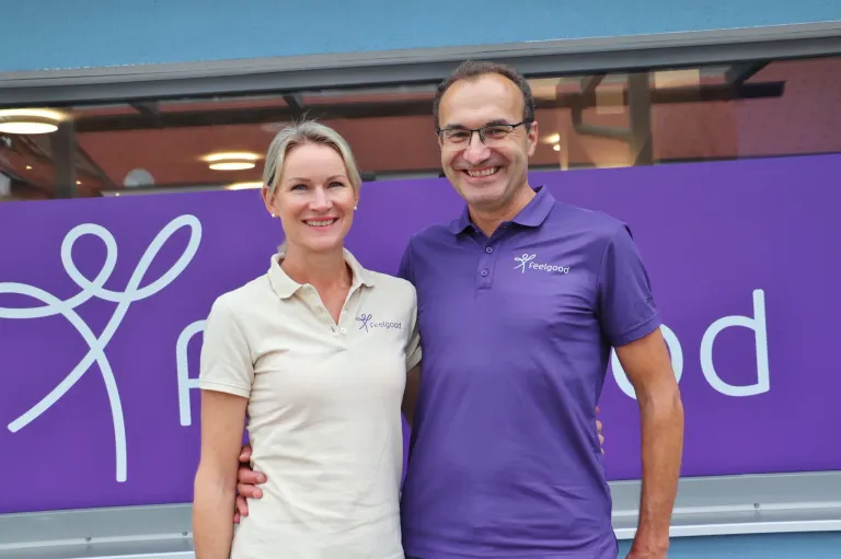 Ein Mann und eine Frau stehen nebeneinander, lächeln und posieren für ein Foto vor einer lila Wand.