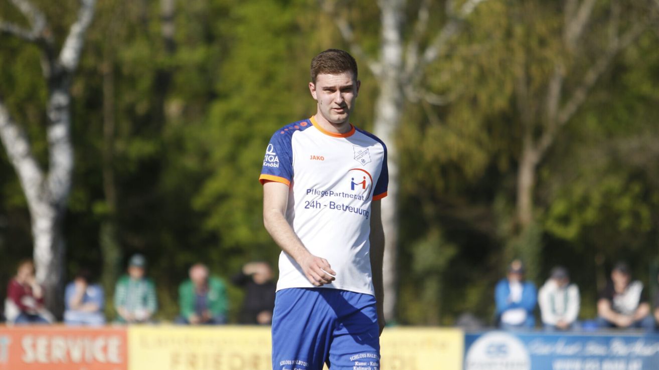 Ein Fußballspieler in einem blau-weißen Trikot steht auf dem Feld. Er schaut zur Seite. Hinter ihm befinden sich verschwommene Figuren von Zuschauern, die das Spiel beobachten.