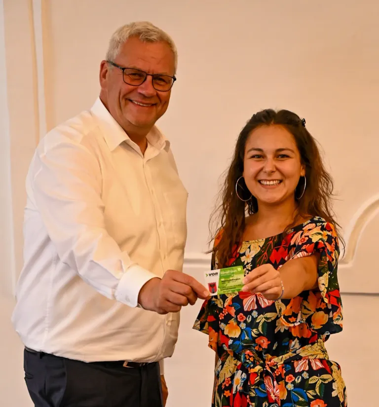 Ein Mann mit Brille überreicht einer jungen Frau eine Karte mit dem Wort 'Vor' darauf. Der Mann lächelt und die Frau schaut auf die Karte und lächelt.