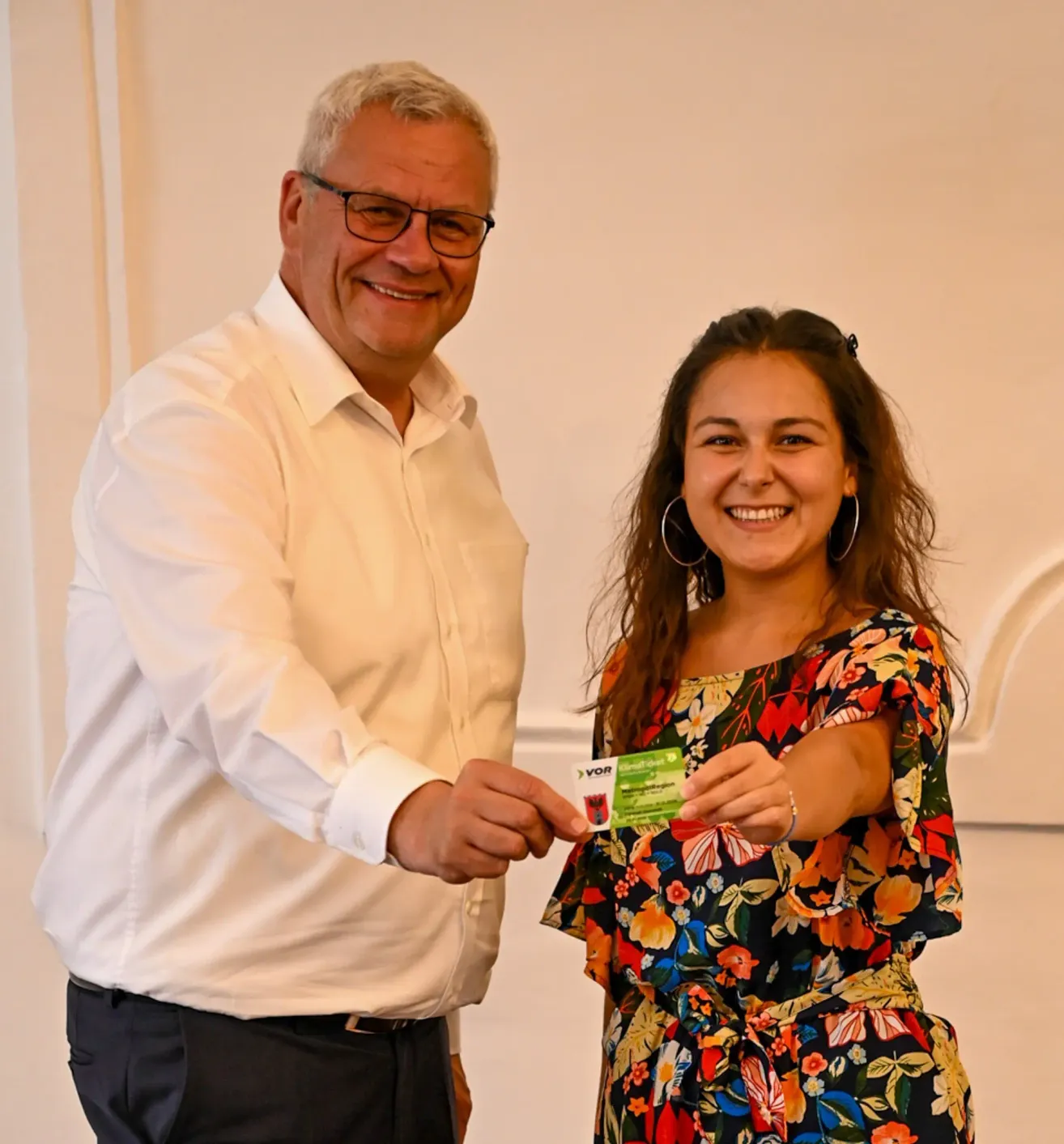 Ein Mann mit Brille überreicht einer jungen Frau eine Karte mit dem Wort 'Vor' darauf. Der Mann lächelt und die Frau schaut auf die Karte und lächelt.