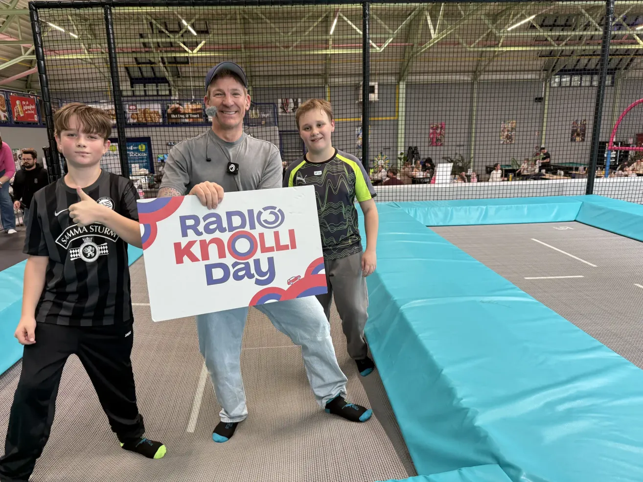Ein Mann und zwei Jungen stehen vor einem Radio Knolli Day Schild in einer Trampolinhalle.