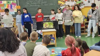Eine Gruppe von Kindern versammelt sich in einem Klassenzimmer. Einige stehen, andere sitzen. Eine Lehrerin ist anwesend und hinter den Kindern befindet sich eine Tafel mit Schrift.