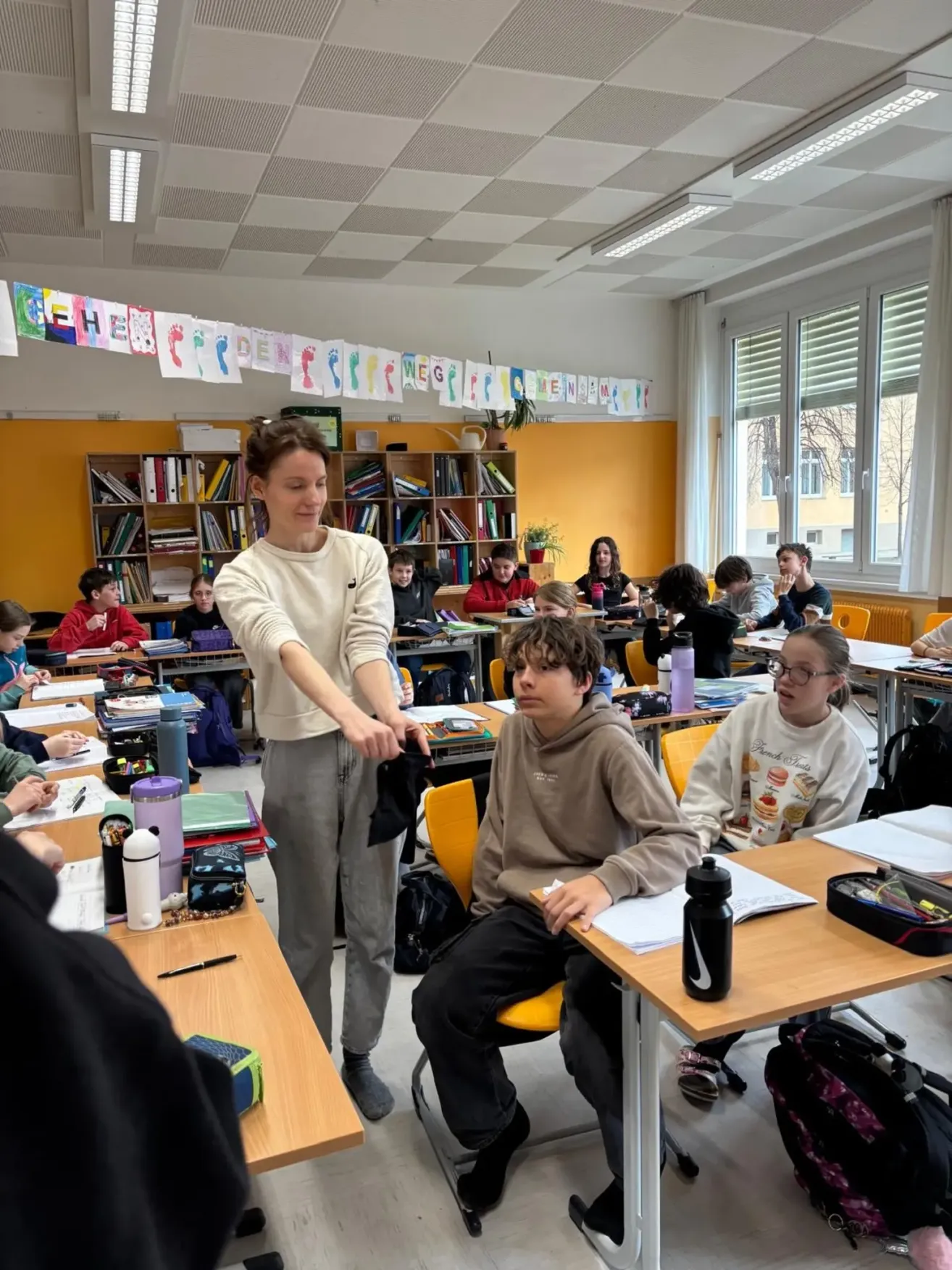 Eine Frau steht in einem Klassenzimmer mit Schülern, die an ihren Schreibtischen sitzen und sich mit Lernaktivitäten beschäftigen. Das Klassenzimmer ist mit bunten Dekorationen und Bücherregalen mit Büchern geschmückt.