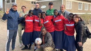 Eine Gruppe von Menschen, einige in roten Trikots, posiert mit einem Fußball vor einem Ziegelgebäude.