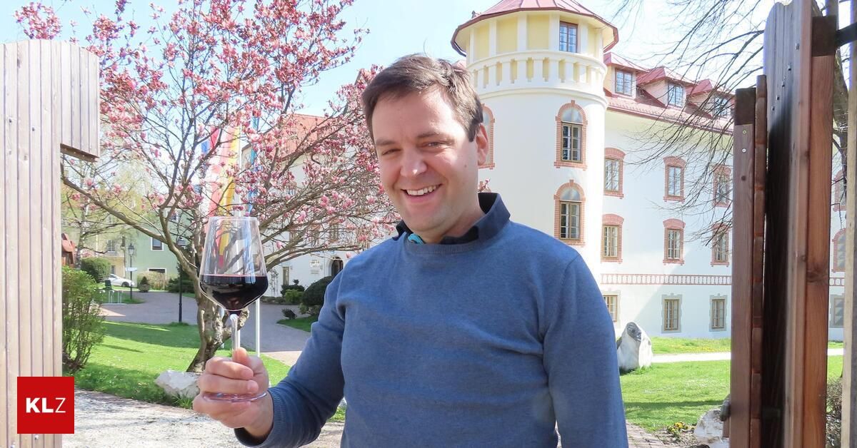 Ein Mann in einem blauen Pullover hält ein Glas Rotwein vor einem historischen Gebäude mit einem rosafarbenen Baum im Hintergrund.