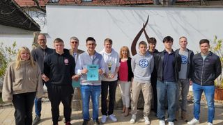 Eine Gruppe junger Leute posiert mit einem Pokal und einem Zertifikat vor einer weißen Wand und einer dekorativen Skulptur.