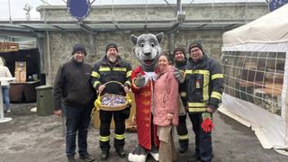 Ein Maskottchen mit einem Körbchen voller Süßigkeiten steht mit vier Feuerwehrleuten, einer hält einen Korb, alle lächeln für die Kamera vor einer Steinmauer.