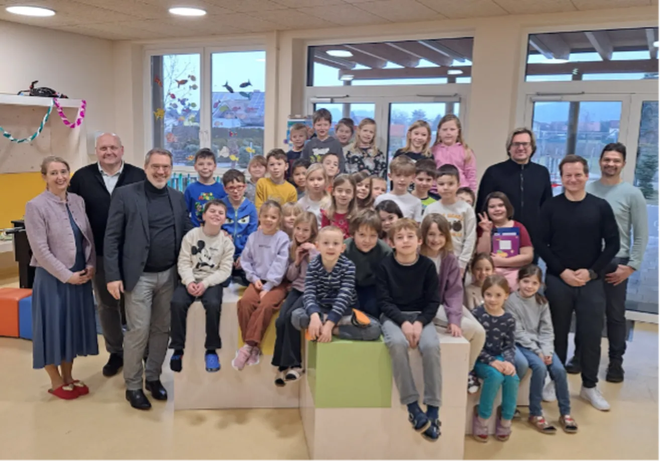 Eine Gruppe von Kindern und Erwachsenen lächelt für ein Foto in einem Klassenzimmer mit Glasfenstern und Fischdekorationen.