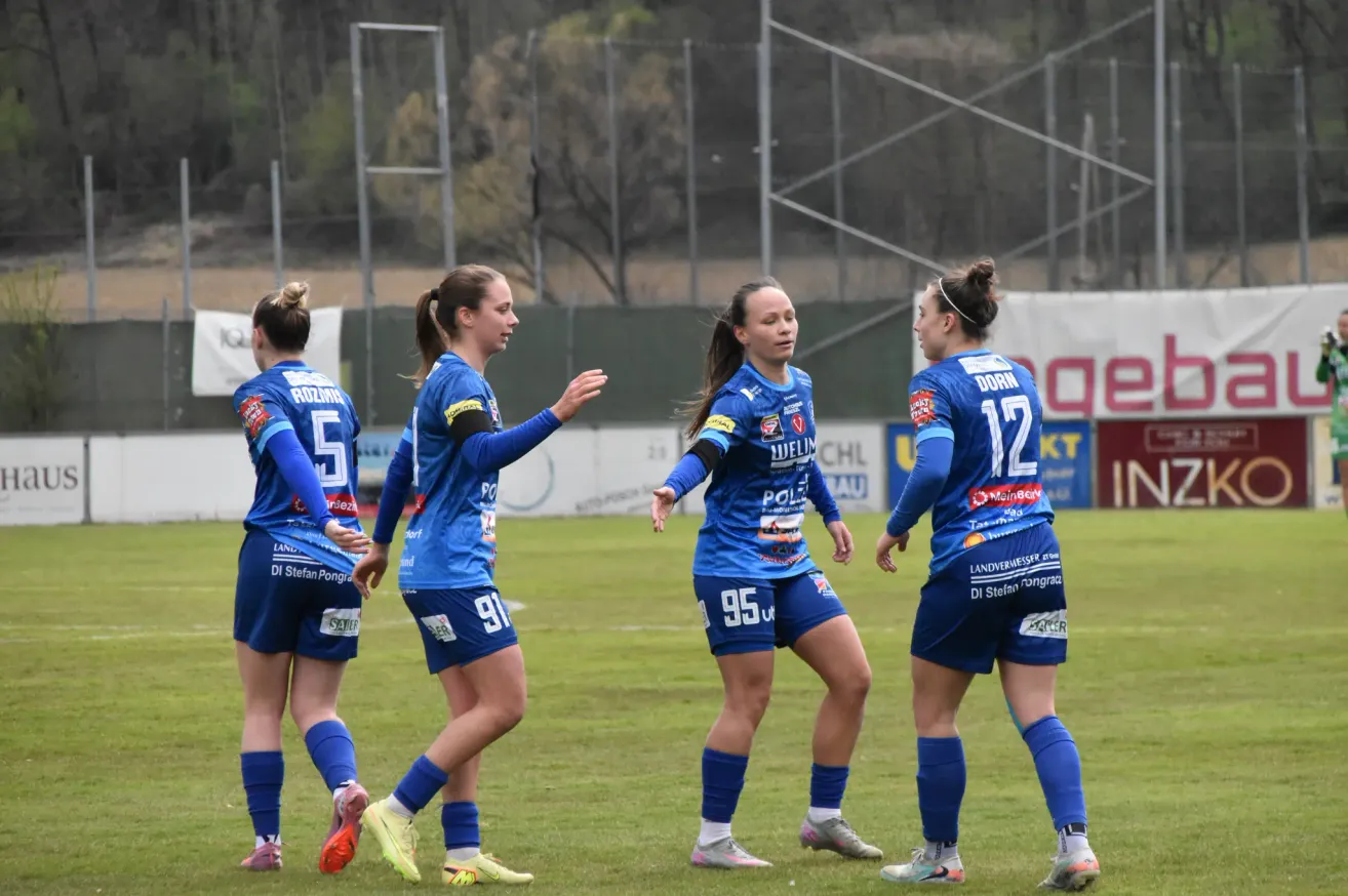 Fünf Fußballspielerinnen in blauen Trikots stehen auf einem Feld. Die Spielerin mit der Nummer 95 schaut die Spielerin mit der Nummer 12 an. Hinter ihnen ist ein verschwommener Blick auf einen Zaun und Bäume.