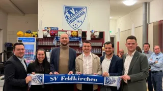 Fünf Männer und eine Frau halten ein blaues Banner mit der Aufschrift 'SV Feldkirchen', möglicherweise zur Feier eines Jubiläums. Dahinter zeigt eine Wand ein Logo und das Jahr 1974.