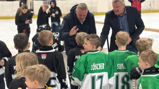 Mehrere Hockeyspieler in Trikots versammeln sich um zwei Männer auf einer Eisbahn. Die Spieler tragen grüne und weiße Trikots mit den Nummern 17 und 18. Der Mann rechts trägt eine Brille.