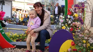 Eine Frau und ein kleines Mädchen lächeln und posieren für ein Foto in einem Park, umgeben von Blumen und einem bunten Objekt. Ein Mann steht im Hintergrund.