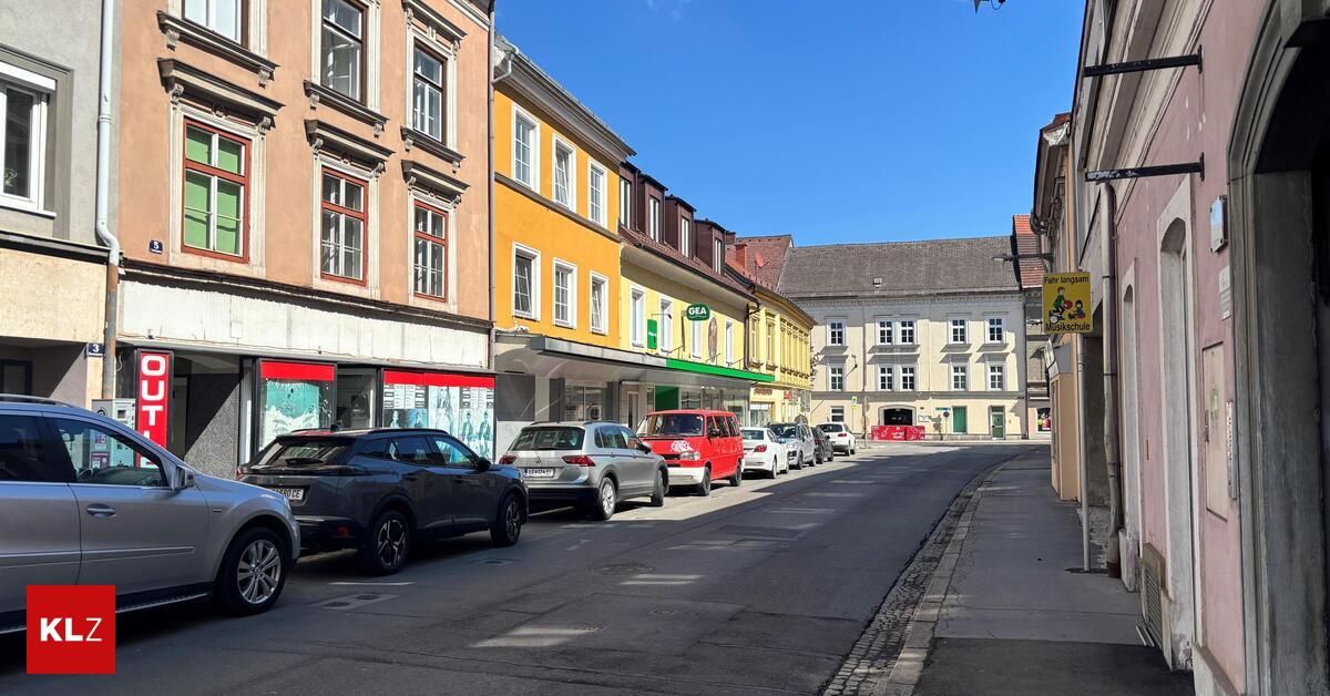 Eine Straße mit geparkten Autos auf einer Seite und Gebäuden mit Geschäften auf der anderen. Die Gebäude haben mehrere Fenster und einen klaren blauen Himmel im Hintergrund.