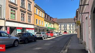 Eine Straße mit geparkten Autos auf einer Seite und Gebäuden mit Geschäften auf der anderen. Die Gebäude haben mehrere Fenster und einen klaren blauen Himmel im Hintergrund.