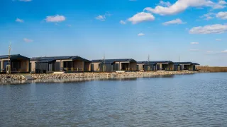 Eine Reihe moderner Hütten mit Solarpanelen auf den Dächern, am Ufer eines Sees unter einem blauen Himmel mit verstreuten Wolken gelegen.