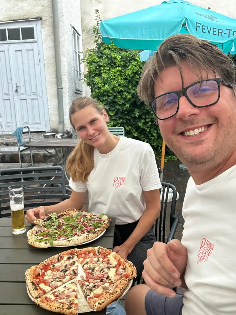 Zwei Personen, ein Mann und eine Frau, sitzen draußen an einem Tisch und lächeln. Vor ihnen stehen zwei große Pizzen. Hinter ihnen befindet sich ein Gebäude mit einer weißen Tür.