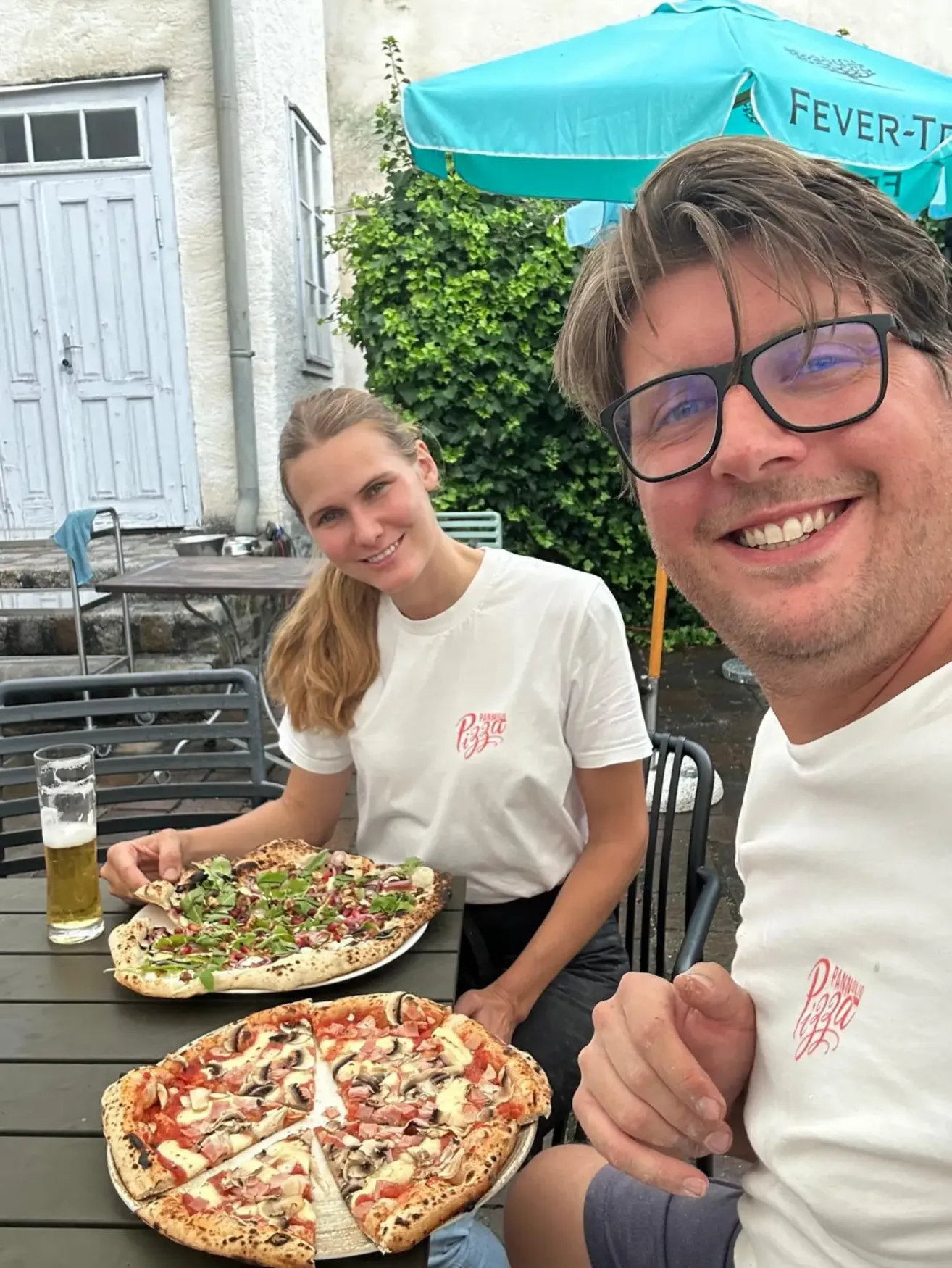Zwei Personen, ein Mann und eine Frau, sitzen draußen an einem Tisch und lächeln. Vor ihnen stehen zwei große Pizzen. Hinter ihnen befindet sich ein Gebäude mit einer weißen Tür.