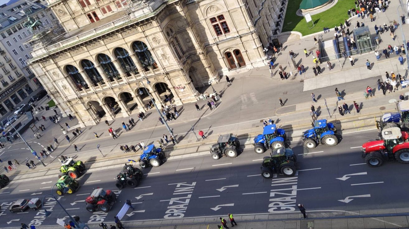 Luftaufnahme einer Straße mit aufgereihten Traktoren, Gebäuden mit vielen Fenstern und Menschen, die auf dem Bürgersteig und im Park spazieren.
