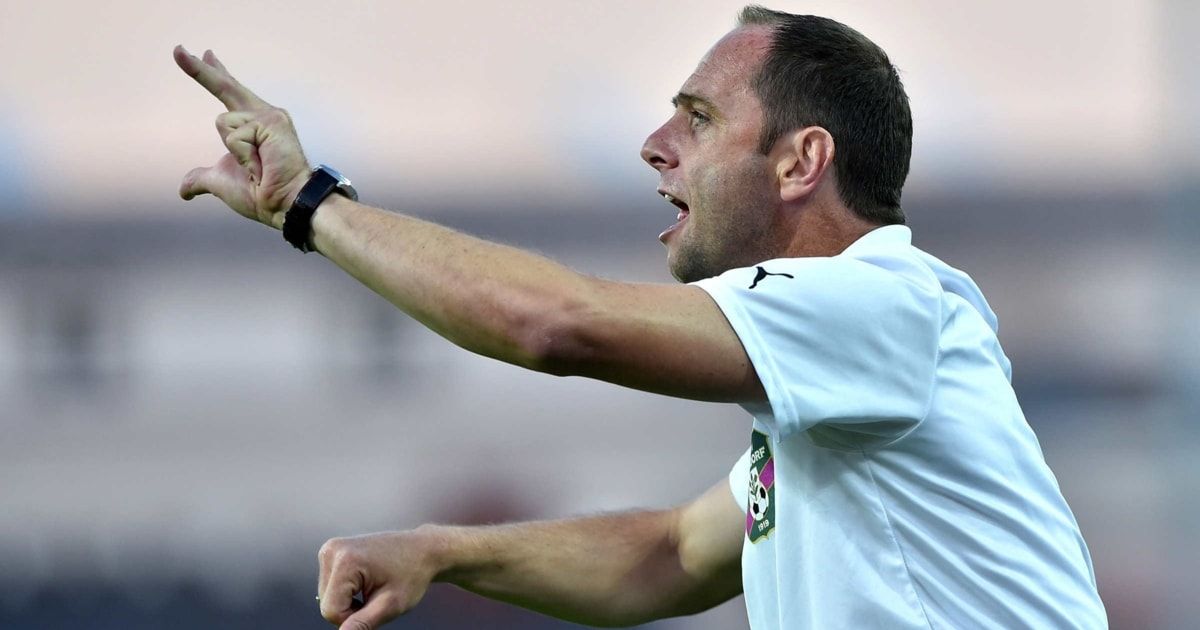 Ein Fußballtrainer in einem weißen Trikot schreit Anweisungen und zeigt auf einem Feld.