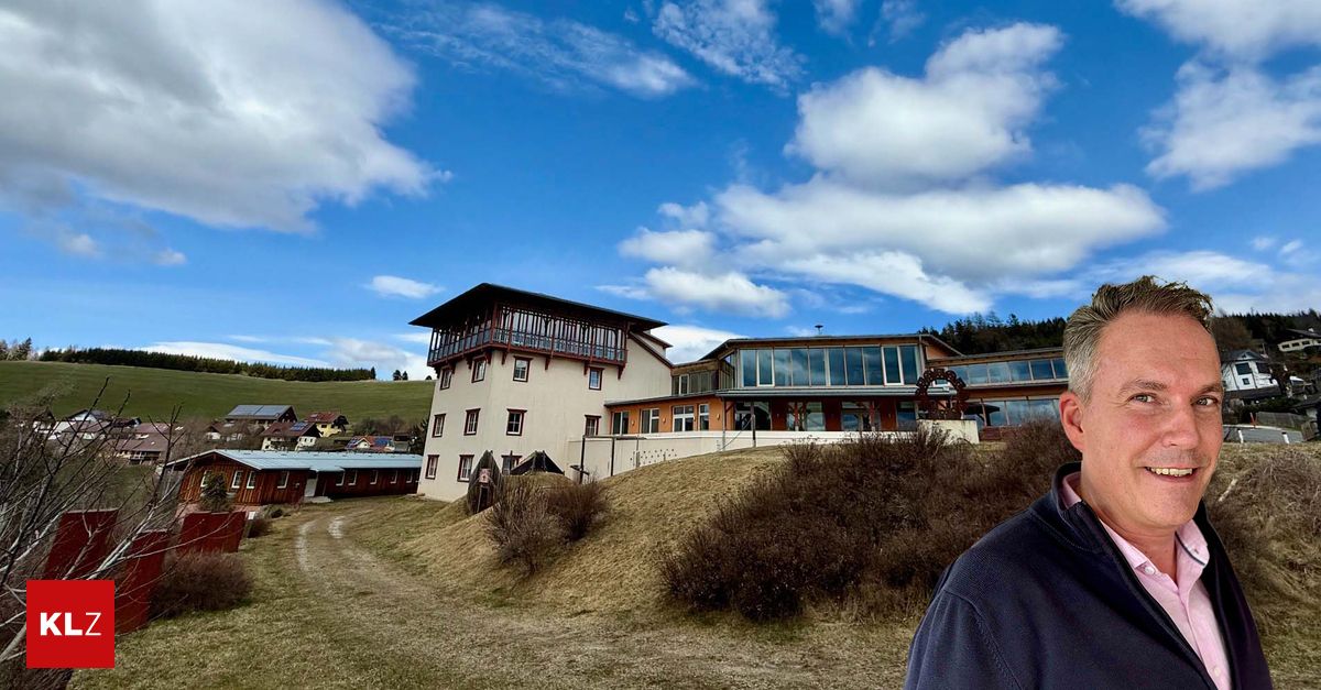 A man stands in front of a large building with many windows and a tower, set against a backdrop of a hill, greenery, and a blue sky with clouds.